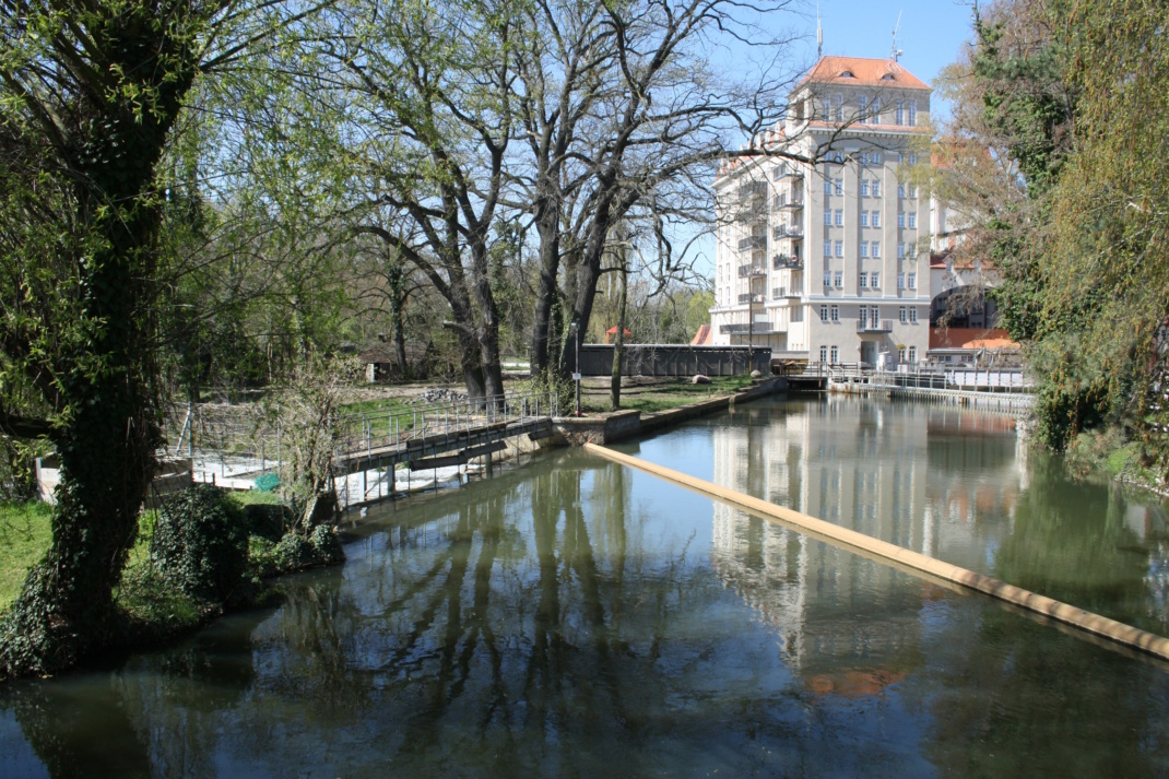 Gewässer mit Gebäude im Hintergrund, Baumbewuchs und Steg.
